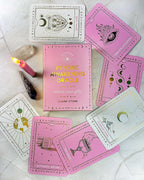A boxed oracle card deck called Psychic Awakening Oracle by Claire Stone is arranged on a light marble-like surface with several illustrated cards fanned around it. The box is pink with gold lettering, and the cards show pastel pink and white designs with gold line art.