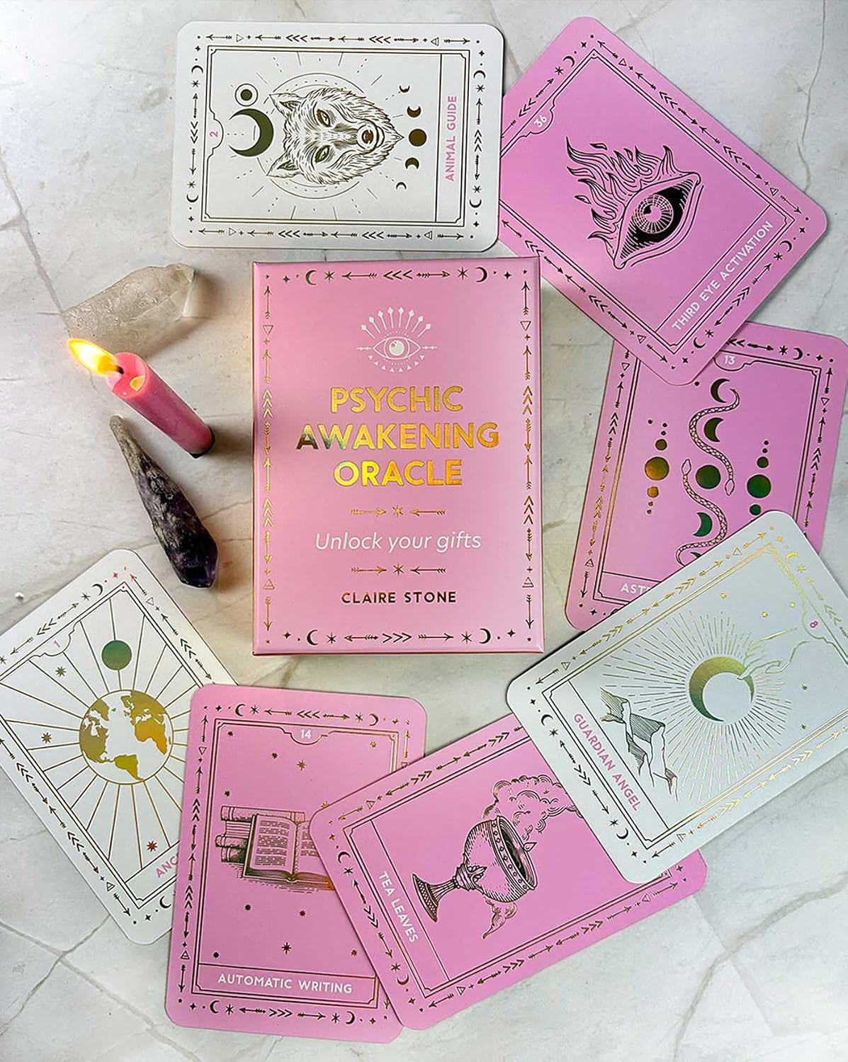 A boxed oracle card deck called Psychic Awakening Oracle by Claire Stone is arranged on a light marble-like surface with several illustrated cards fanned around it. The box is pink with gold lettering, and the cards show pastel pink and white designs with gold line art.