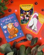 A styled flat-lay of a 'Get Wisdom Oracle' card deck with two illustrated cat oracle cards and the book cover, arranged on a bright orange background with decorative leaves, paw prints, and crystals.