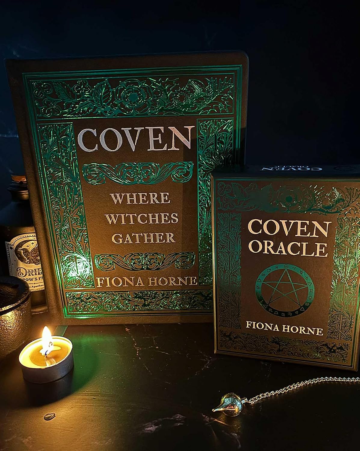 Two books titled 'Coven' and 'Coven Oracle' by Fiona Horne on a dark surface with a lit candle.