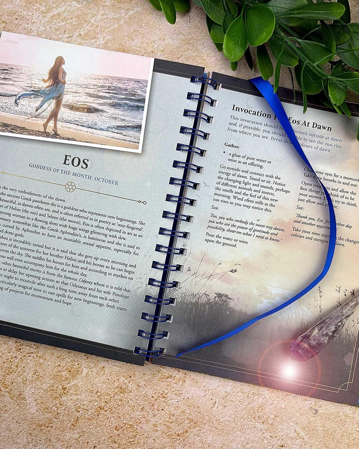 An open spiral-bound journal or prayer book laid on a beige stone surface, with printed text pages visible, a small beach photo insert on the left page, and a bright blue ribbon draped across the right page. Green leaves and a small crystal pendant are placed around it as styled props.