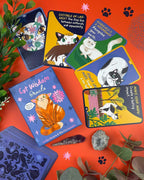 A Cat Wisdom Oracle card deck and several illustrated cards are arranged on a bright orange surface with decorative leaves, crystals, and paw-print graphics. The box and cards feature colorful cat illustrations and text prompts related to cats.
