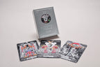 A boxed tarot card deck standing upright with three illustrated tarot cards spread in front on a plain light background. The box is metallic silver with a circular cutout showing a hand illustration, and the cards feature black, white, gray, and small red/orange accents in the artwork.