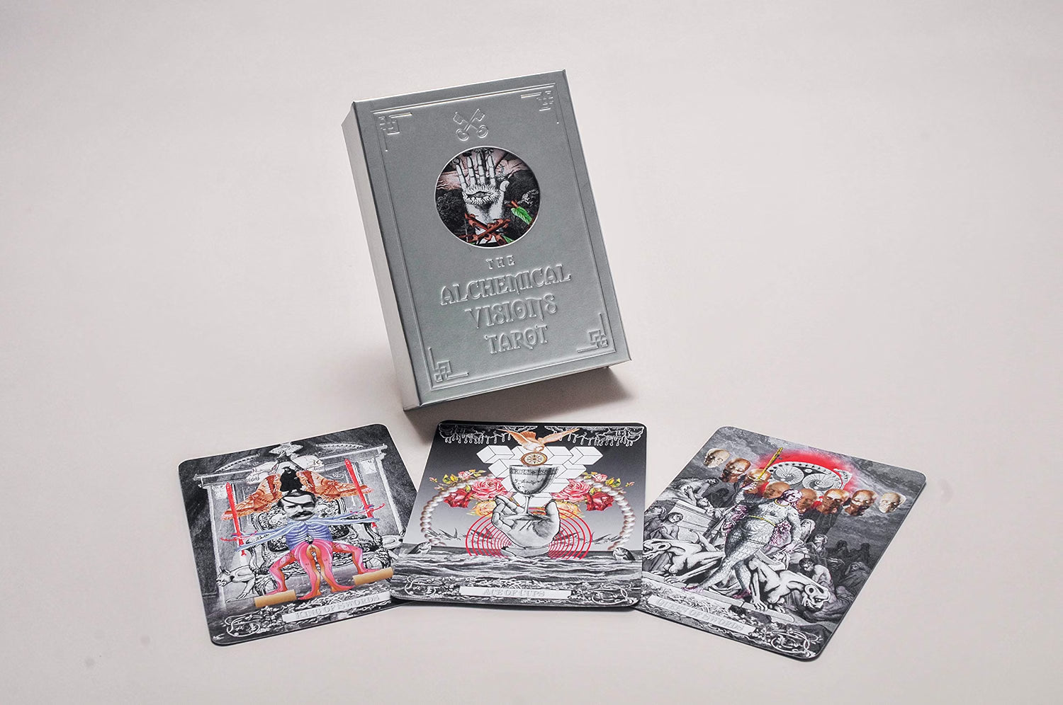 A boxed tarot card deck standing upright with three illustrated tarot cards spread in front on a plain light background. The box is metallic silver with a circular cutout showing a hand illustration, and the cards feature black, white, gray, and small red/orange accents in the artwork.