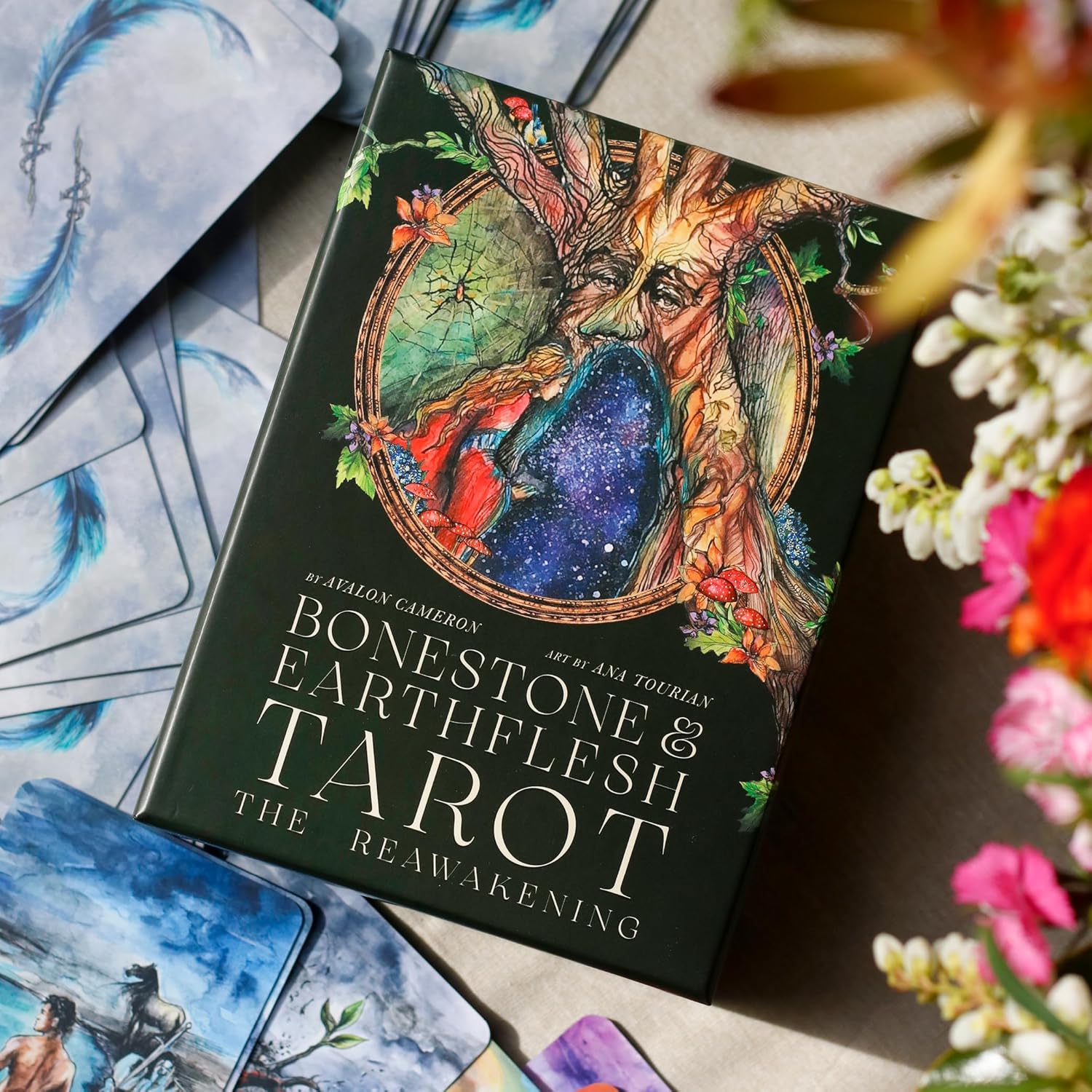 Book titled 'Bonestone & Earthflesh Tarot' surrounded by tarot cards and flowers on a wooden surface.
