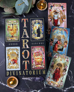 A tarot deck set arranged on a dark marble surface, including a black box labeled 'TAROT DIVINATORIUM' by André Sanchez, several illustrated tarot cards, candles, green leaves, and a crystal.
