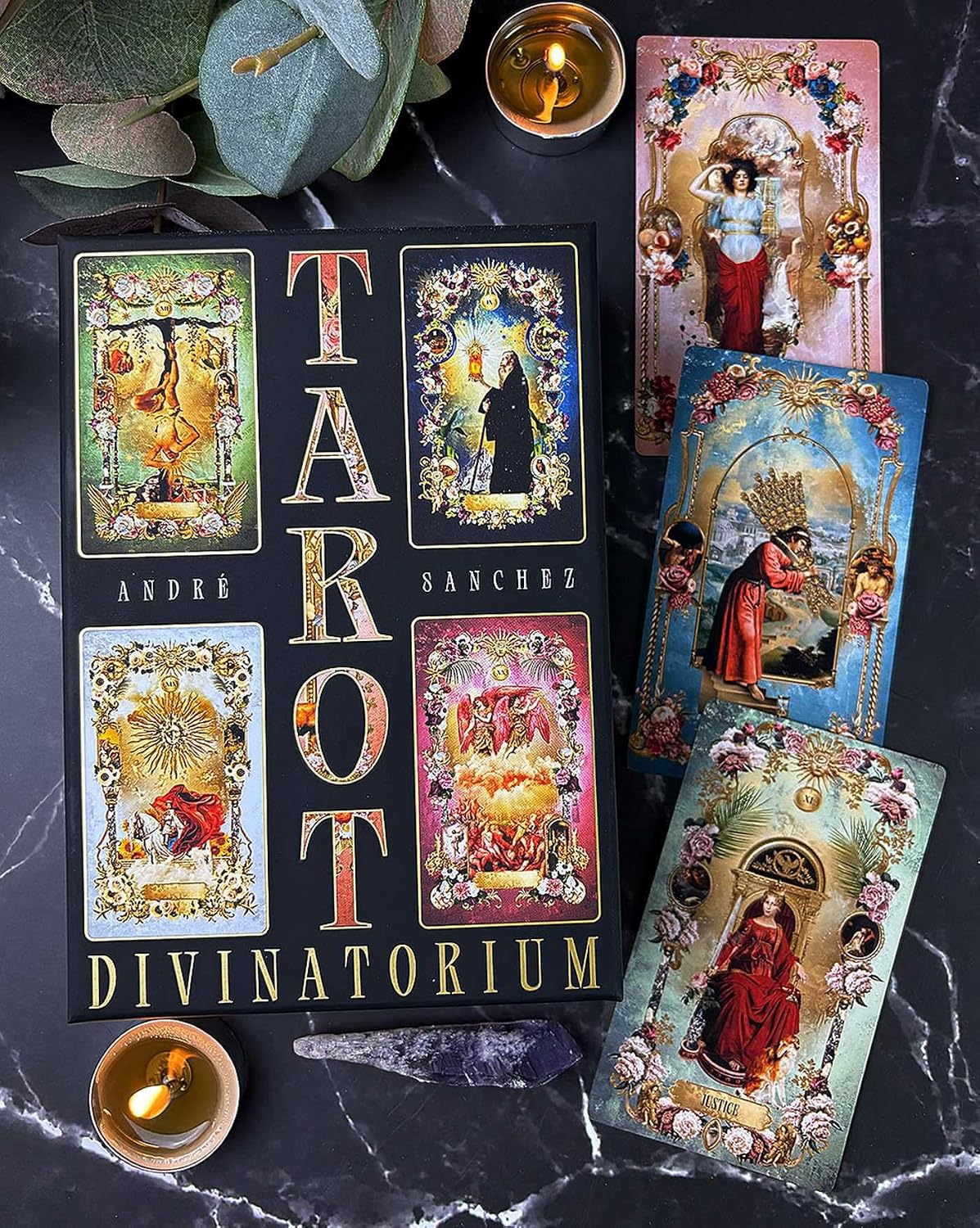 A tarot deck set arranged on a dark marble surface, including a black box labeled 'TAROT DIVINATORIUM' by André Sanchez, several illustrated tarot cards, candles, green leaves, and a crystal.