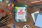 Guided Art Therapy Card Deck surrounded by art supplies on a wooden surface
