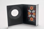 An open black presentation box or album-style package on a light gray studio background, showing printed artwork and text on both interior panels.