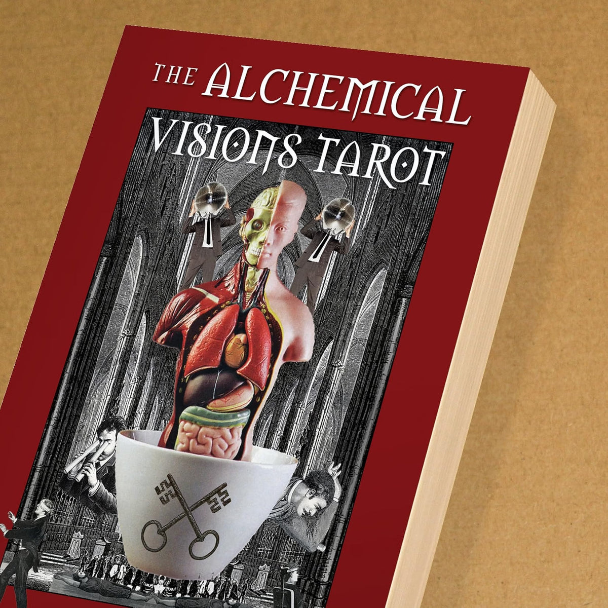 The Alchemical Visions Tarot deck box shown at an angle on a textured tan background. The box features a deep red cover with white gothic-style title text and a central surreal illustration of a human torso emerging from a white cup with keys, set against a black-and-white gothic background.