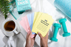 Person holding exercise cards with a cup of tea, dumbbells, and a book on a white surface.