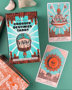 A styled flat-lay of the 'Crossed Destinies Tarot' deck by Bobby Green, showing the illustrated book cover and several tarot cards on a teal surface with small stones nearby.