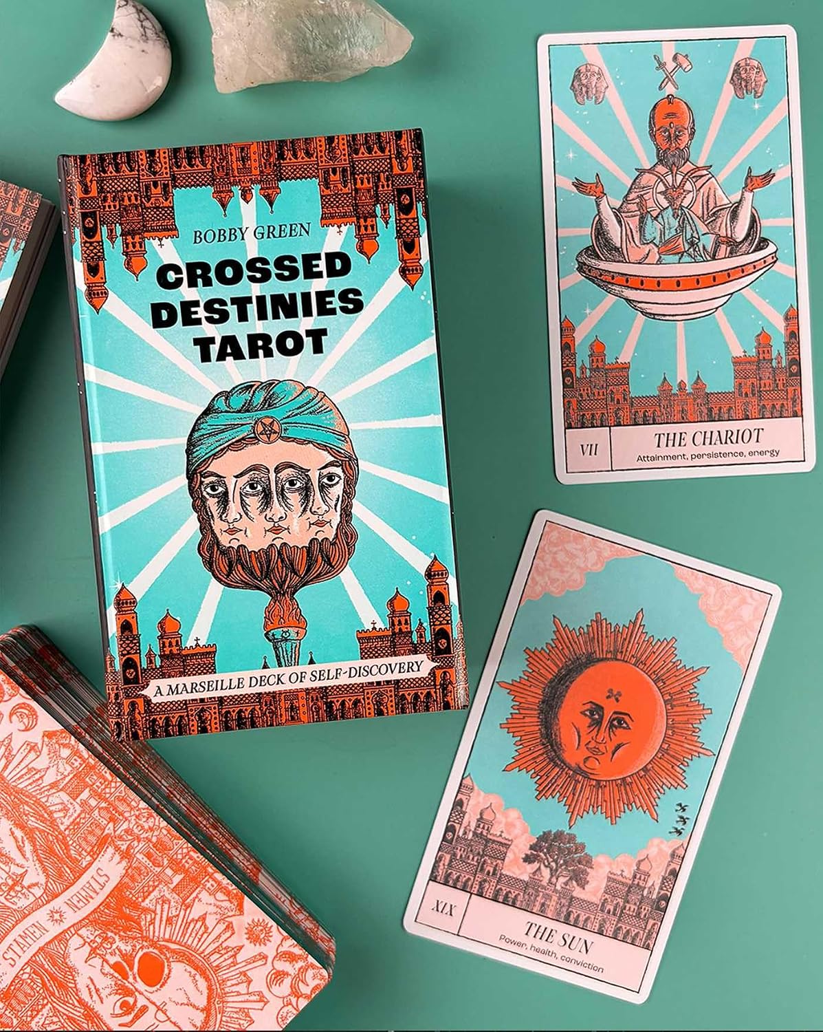 A styled flat-lay of the 'Crossed Destinies Tarot' deck by Bobby Green, showing the illustrated book cover and several tarot cards on a teal surface with small stones nearby.