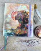 A 2027 Gratitude Diary & Daily Planner with a colorful, artistic cover featuring a woman's profile and celestial elements, styled on a textured stone surface with a feather, crystal, and paintbrush.