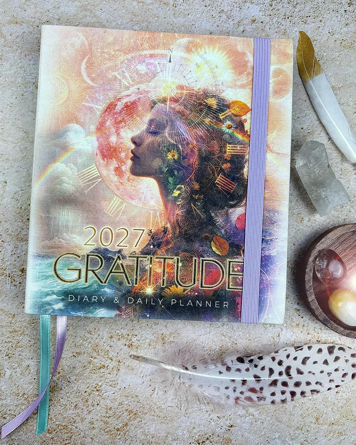 A 2027 Gratitude Diary & Daily Planner with a colorful, artistic cover featuring a woman's profile and celestial elements, styled on a textured stone surface with a feather, crystal, and paintbrush.