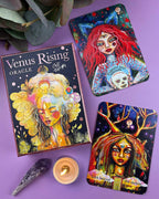 Venus Rising Oracle card deck box and two illustrated oracle cards arranged on a lavender background with a candle, crystal, and green leaves as styled props.