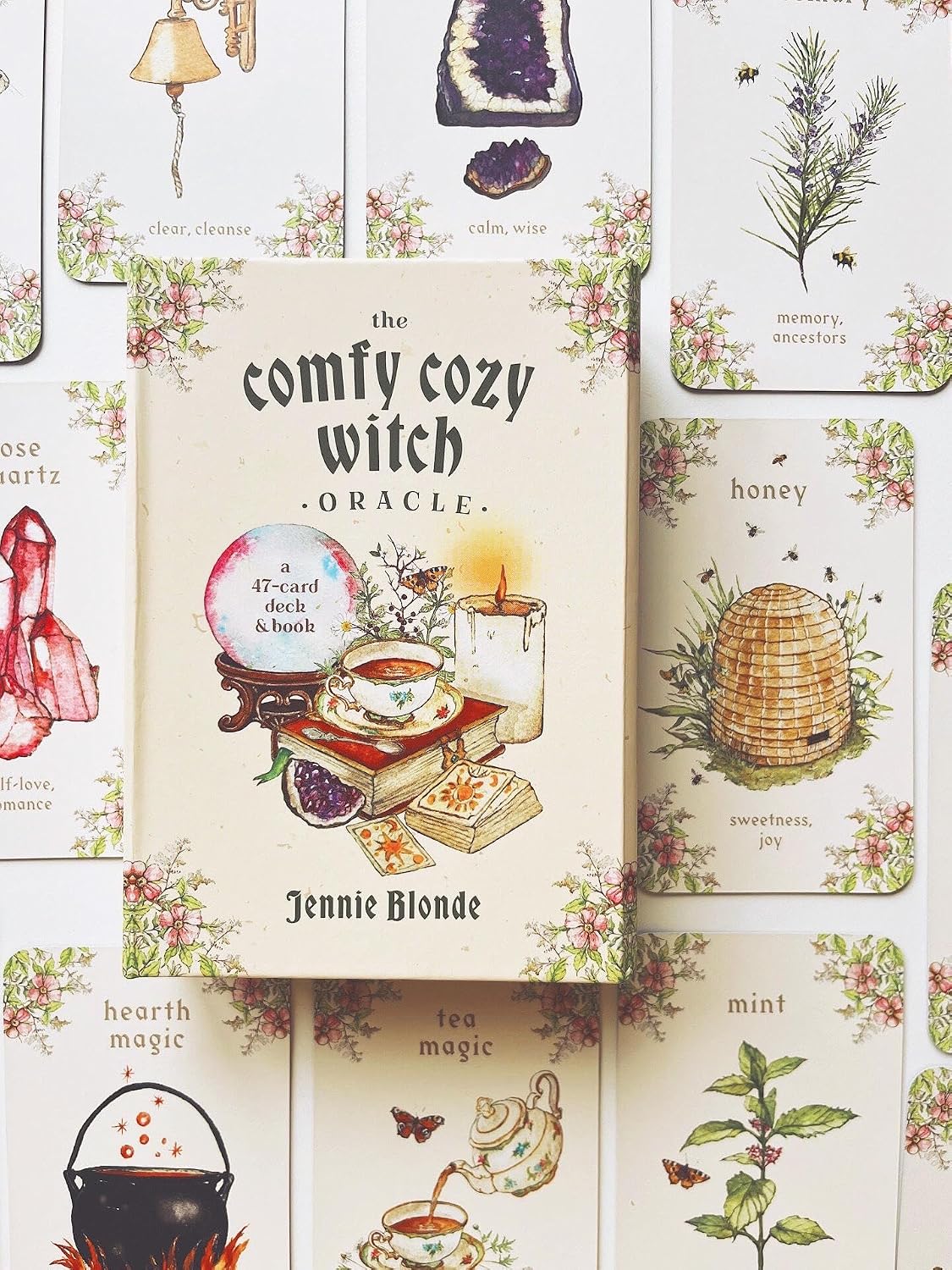The front cover of 'The Comfy Cozy Witch Oracle' deck box is centered, surrounded by illustrated oracle cards with vintage-style drawings of crystals, herbs, a beehive, a kettle, and tea accessories on a warm cream background.