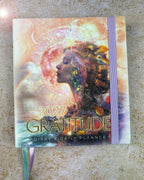 A 2027 Gratitude Diary & Daily Planner with a colorful cosmic collage cover, featuring a profile of a woman with a moon and rainbow motif. The planner has a lavender elastic closure band and a matching ribbon bookmark, photographed on a textured beige stone surface.