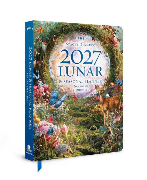 A hardcover journal titled '2027 Lunar & Seasonal Planner' by Stacey Demarco, featuring a highly detailed, colorful nature illustration on the cover with a deer, hedgehog, flowers, and a blue butterfly. The book is shown at an angle on a clean white background.