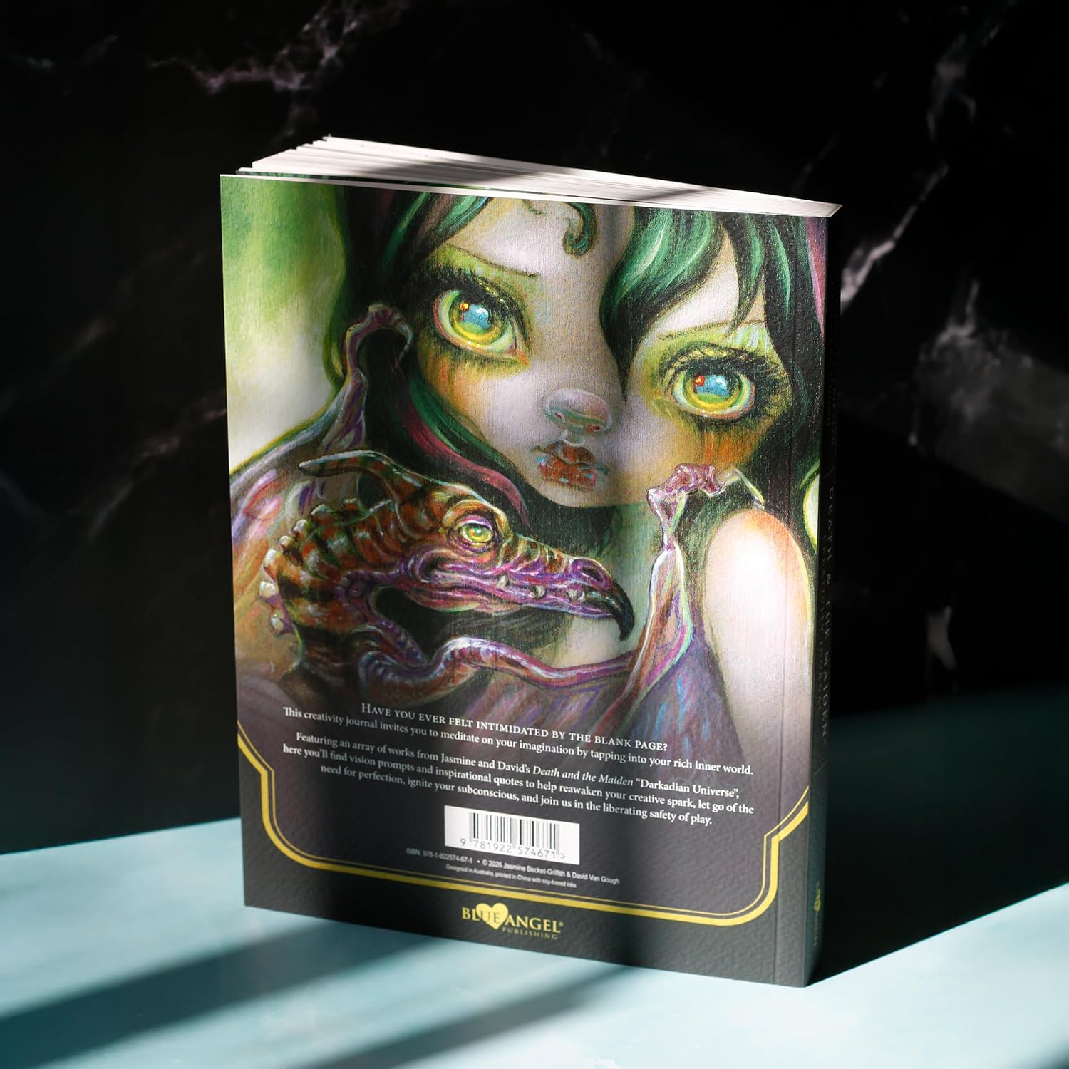 A hardcover art book titled 'The Art of Anger' by D'Angelo, standing upright on a light blue surface against a dark, marbled background. The cover features a highly detailed, colorful illustration of a woman with green hair and glowing eyes holding a dragon.
