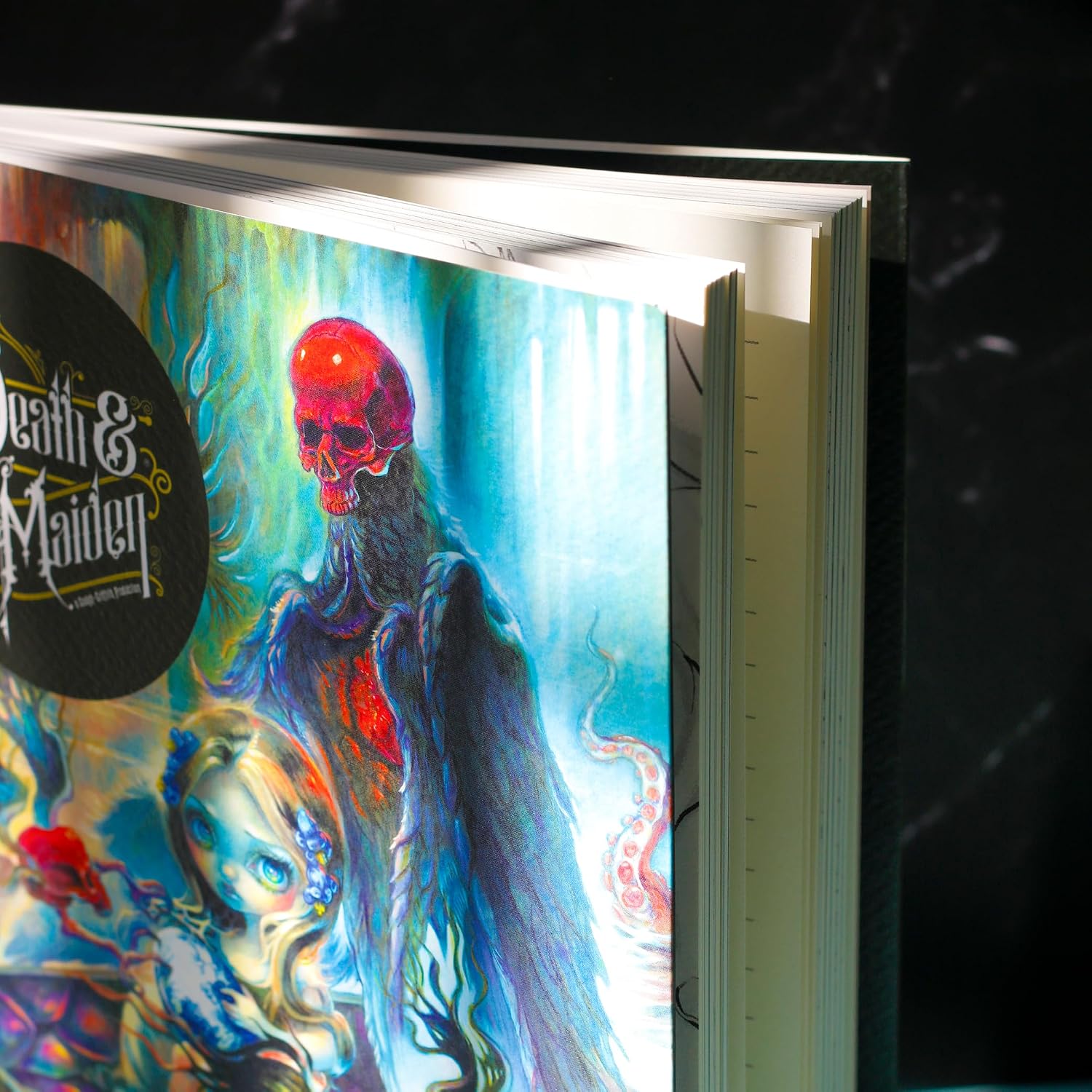 A close-up of a hardcover book or album cover featuring vivid fantasy artwork with a red skull-like figure, blue glowing background, and a black circular logo reading 'Death & Maiden.' The product is photographed at an angle against a dark marble-like surface.
