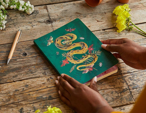 A teal green hardcover notebook with a detailed illustration of a golden snake, butterflies, and leaves, held by two hands on a rustic wooden table with flowers and a pen.
