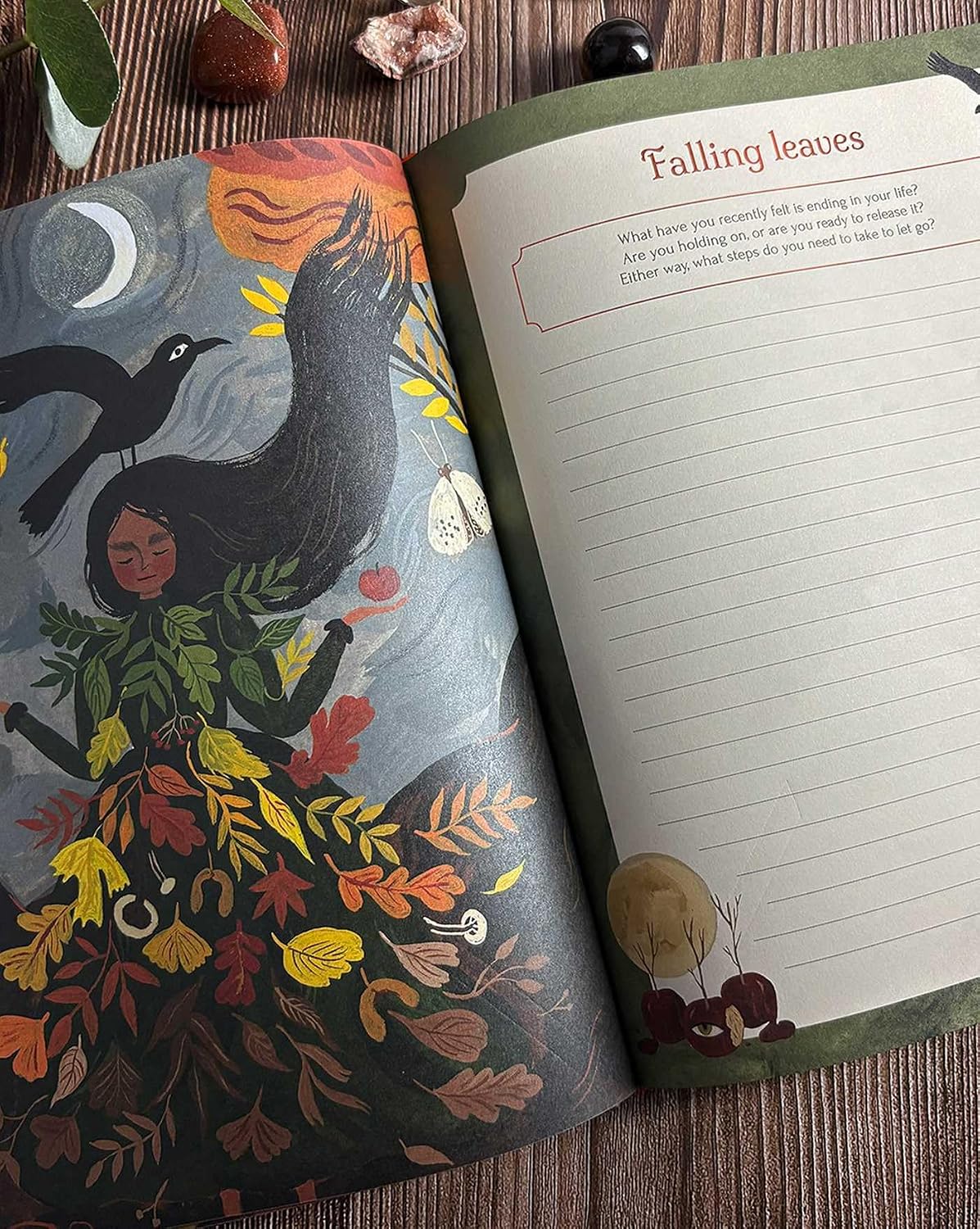 An open journal or notebook spread on a wooden surface, showing a dark fantasy-style illustration on the left page with a raven, moon, and autumn leaves, and a lined writing page titled 'Falling leaves' on the right.