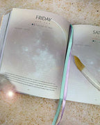 An open journal or planner spread on a speckled beige countertop, showing pages titled Friday and Saturday with a pastel ribbon bookmark and a gold pen resting on the right page.