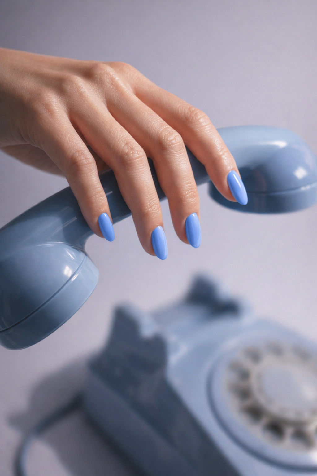 Hand with blue nail polish holding a blue rotary phone on a light gray background