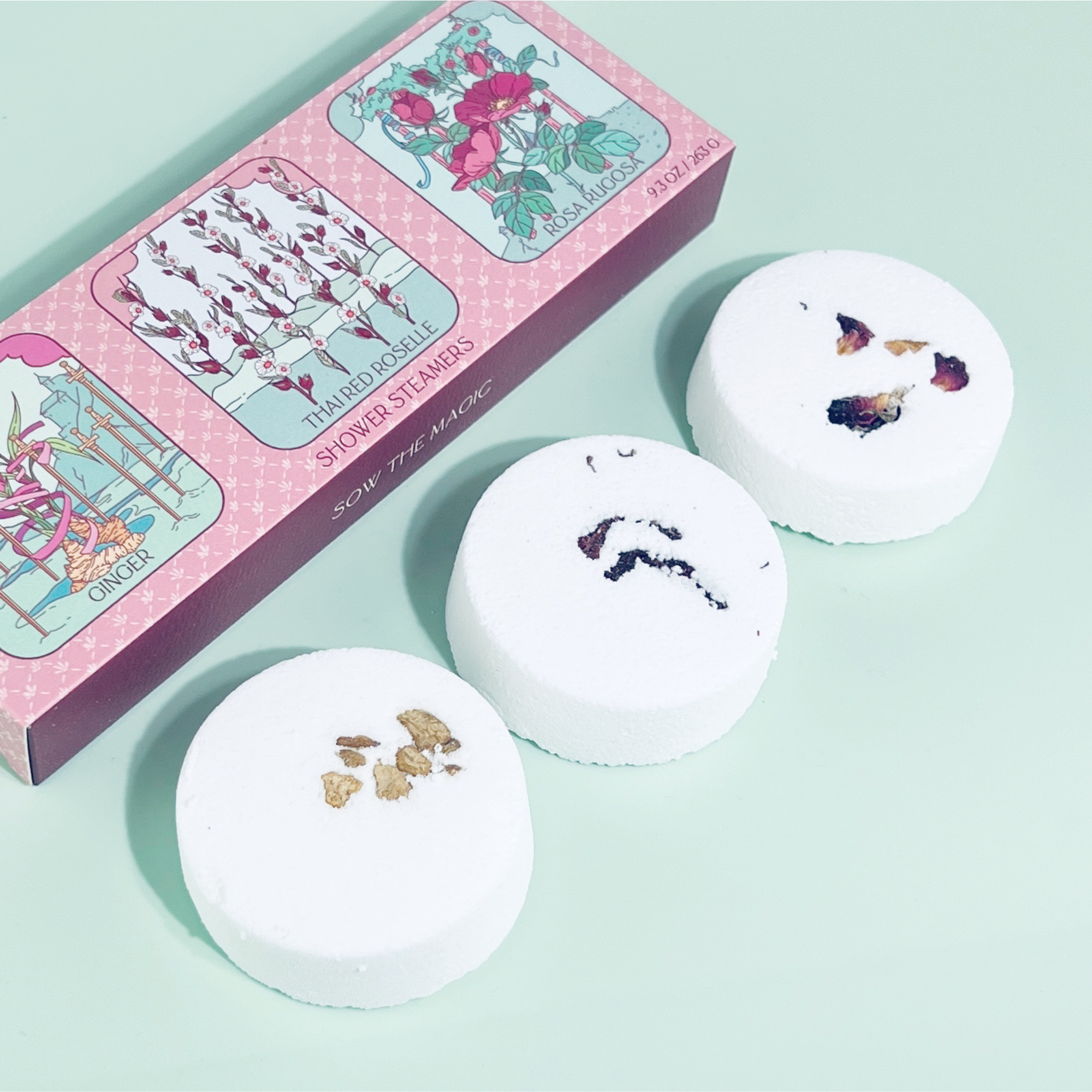 Three white bath bombs with floral designs on a light green background, next to a pink box with illustrations.