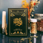 The Cosma Visions Oracle book set by James R. Eads, featuring a black cover with gold lettering and illustration, styled on a dark reflective tabletop with vintage keys, dried flowers, and glass jars in the background.