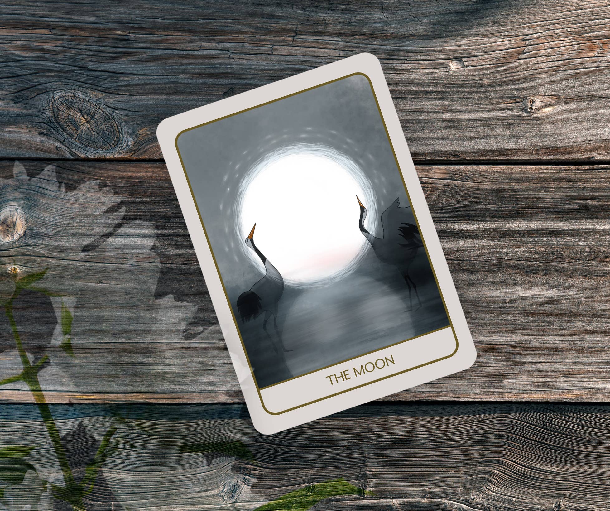 A tarot card titled 'THE MOON' lies diagonally on a rustic weathered wood background. The card features a white border with gold lines and an illustration of two silhouetted cranes facing a bright full moon.