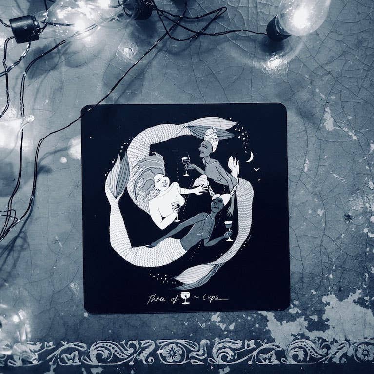 Decorative coaster with a black and white illustration of mermaids on a textured surface with fairy lights.