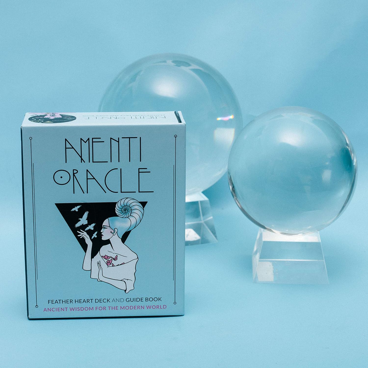 Amentii Oracle card deck box in front of two clear crystal spheres on acrylic stands, photographed on a soft light blue background.