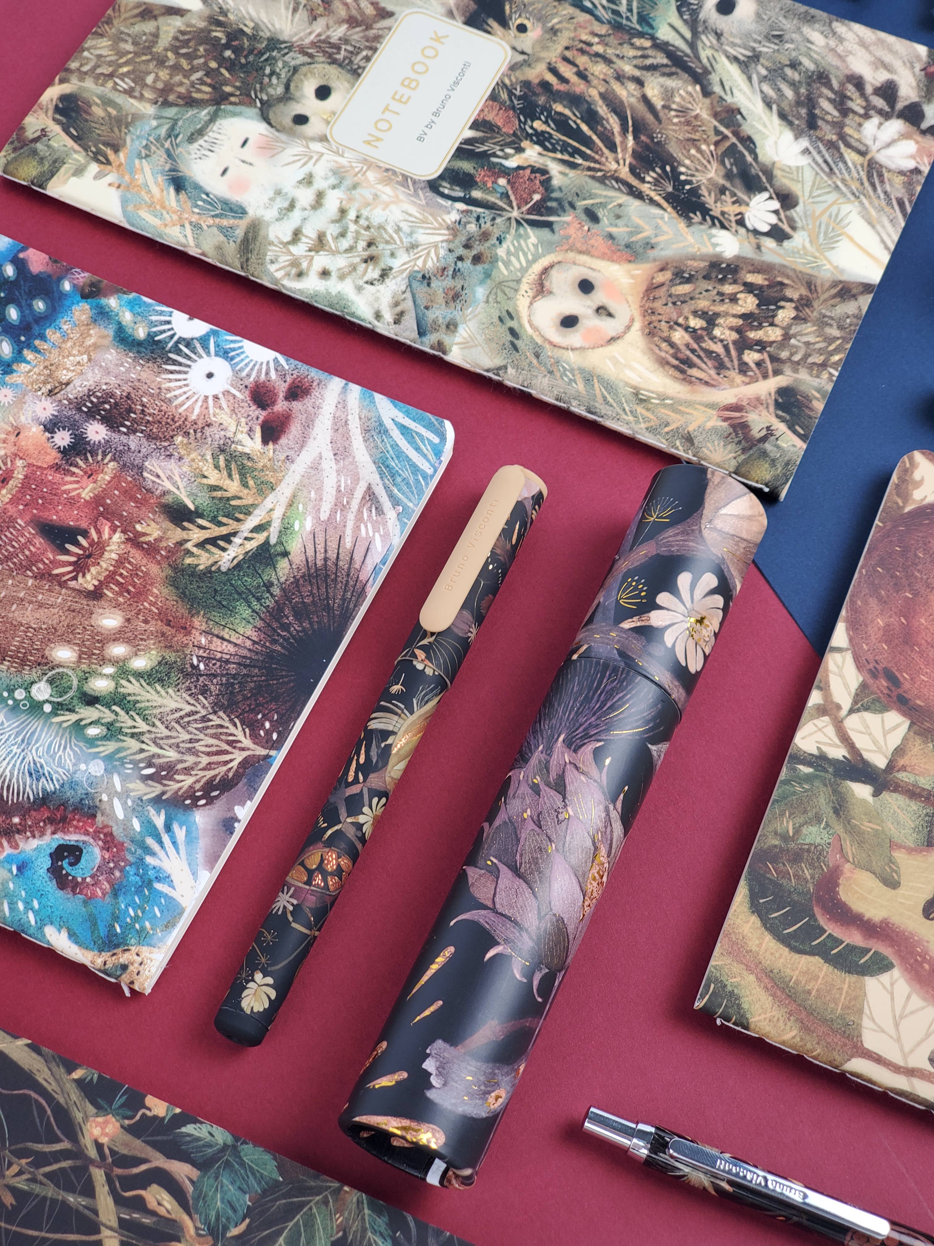 Collection of notebooks and pens with nature-themed designs on a red surface