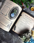 An open vintage-style book spread showing aged parchment pages with text and an elderflower illustration, styled on a dark textured surface with dried flowers, greenery, and a small candle.