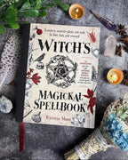 Book titled 'Witch's Magickal Spellbook' surrounded by mystical items on a dark surface.