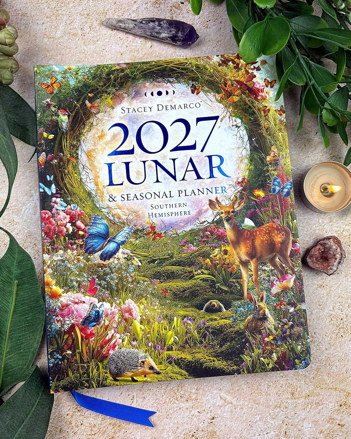 A 2027 Lunar & Seasonal Planner by Stacey Demarco lies flat on a textured beige surface, surrounded by decorative natural props including green leaves, crystals, a small candle, and a blue ribbon bookmark. The cover art is highly detailed and colorful, featuring a forest scene with a deer, hedgehog, butterflies, and a large floral wreath framing the title.