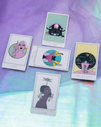 Five illustrated tarot-style cards arranged on iridescent purple and blue fabric. The cards feature stylized portraits and abstract artwork with text at the bottom, including 'I AM LOV', 'I SEE THE BEAUTY IN YOU', 'I AM NOT WEARY', 'I AM NOT WIZARD', and 'INTIMATE THE FULL LIFE IS A RIDE'.