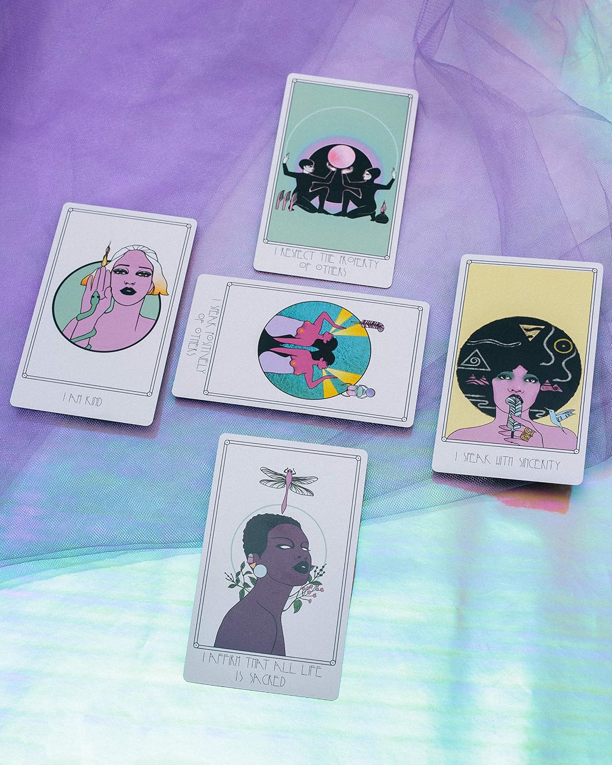 Five illustrated tarot-style cards arranged on iridescent purple and blue fabric. The cards feature stylized portraits and abstract artwork with text at the bottom, including 'I AM LOV', 'I SEE THE BEAUTY IN YOU', 'I AM NOT WEARY', 'I AM NOT WIZARD', and 'INTIMATE THE FULL LIFE IS A RIDE'.