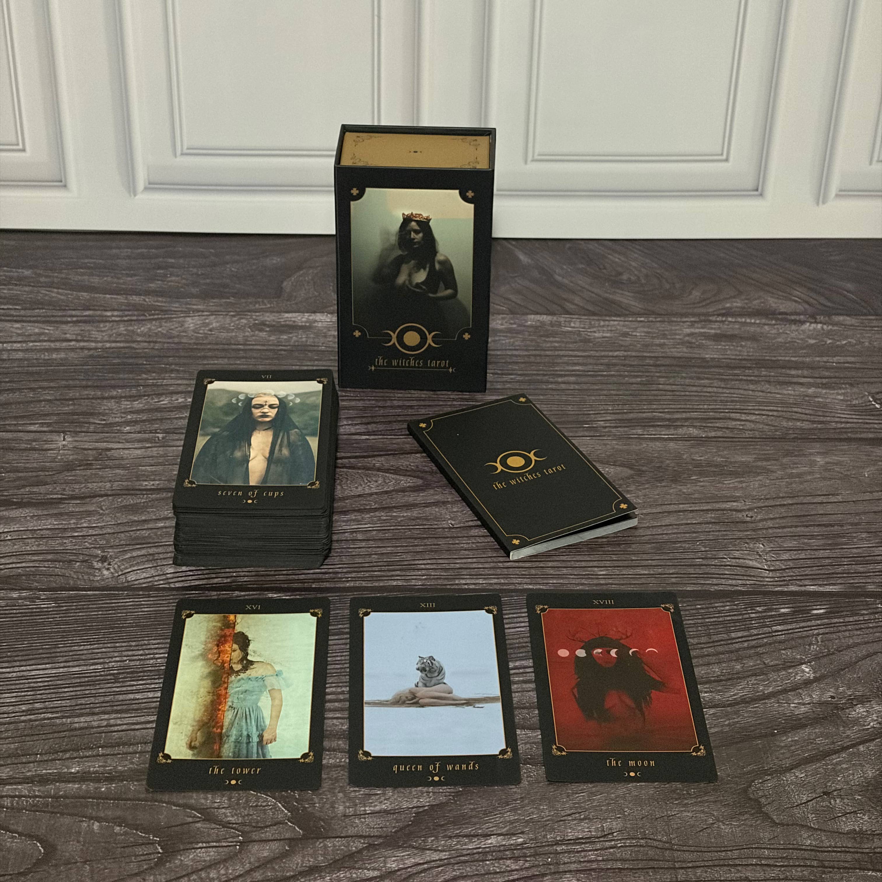 A boxed tarot deck labeled 'The Beast Within' stands upright on a dark wood floor, with several cards fanned out in front. The packaging and cards are black with gold text and border details, and the visible card art includes dark, moody illustrations.