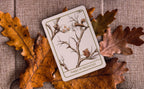 A tarot card labeled 'THREE OF PENTACLES' with an illustration of branches, acorns, and a squirrel, placed on autumn oak leaves over a burlap fabric background.