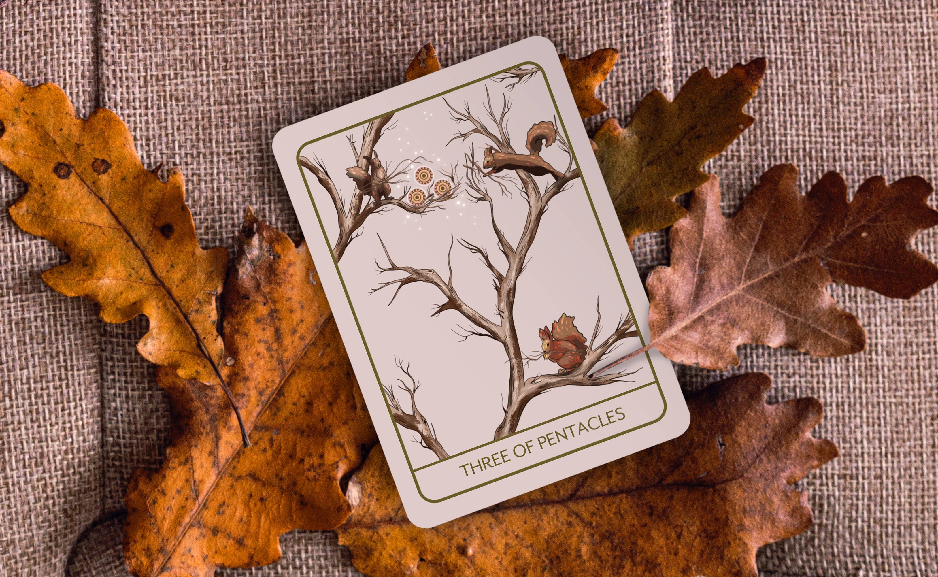 A tarot card labeled 'THREE OF PENTACLES' with an illustration of branches, acorns, and a squirrel, placed on autumn oak leaves over a burlap fabric background.