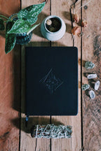 Black notebook with embossed design on a wooden surface with plants and crystals.