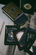 A deck of illustrated tarot cards with ornate black and gold packaging, shown with several cards fanned out on a reflective surface.