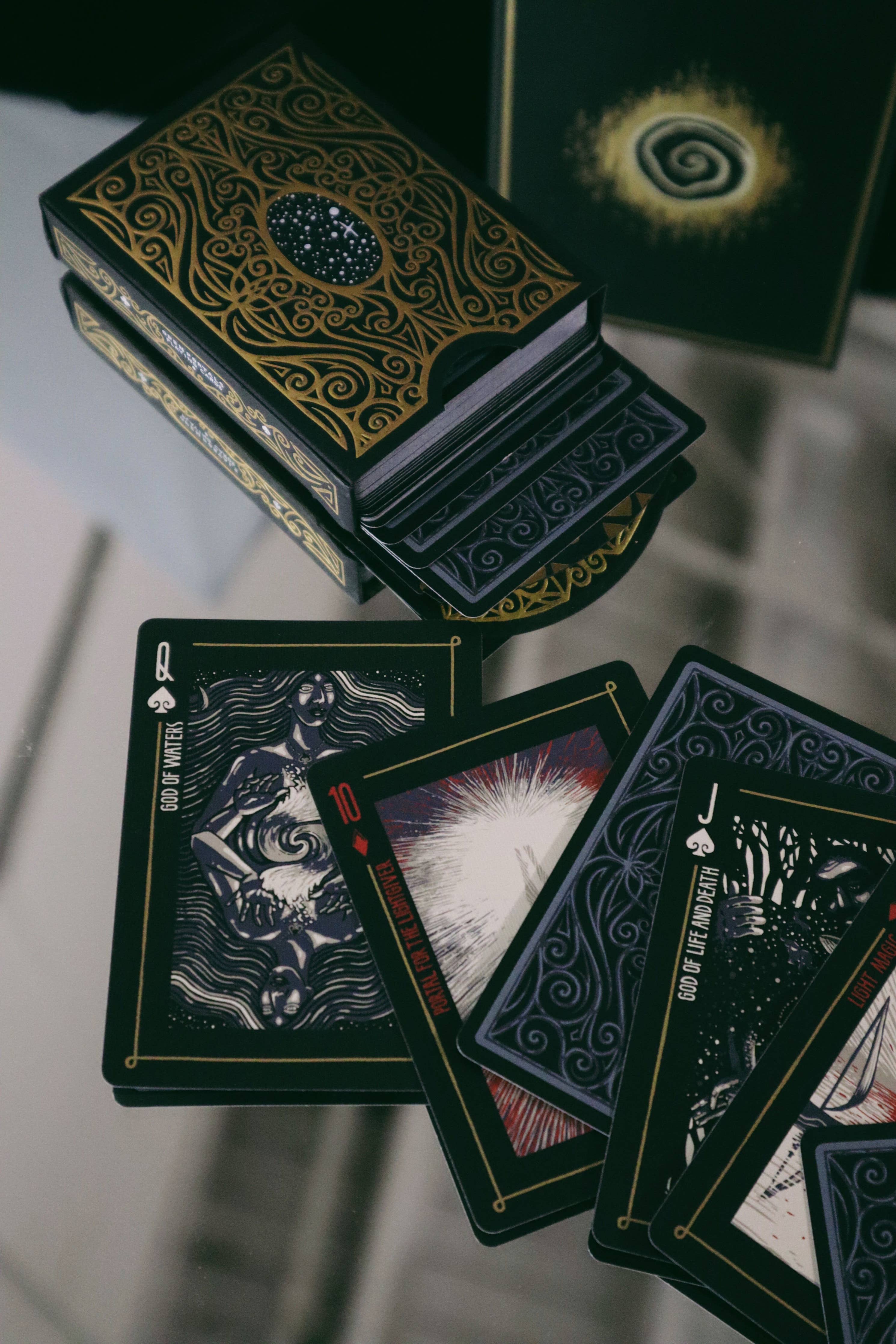 A deck of illustrated tarot cards with ornate black and gold packaging, shown with several cards fanned out on a reflective surface.