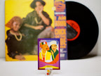 Vinyl record with a colorful cover featuring two people, a smaller card with a similar design, and a yellow album cover in the background.