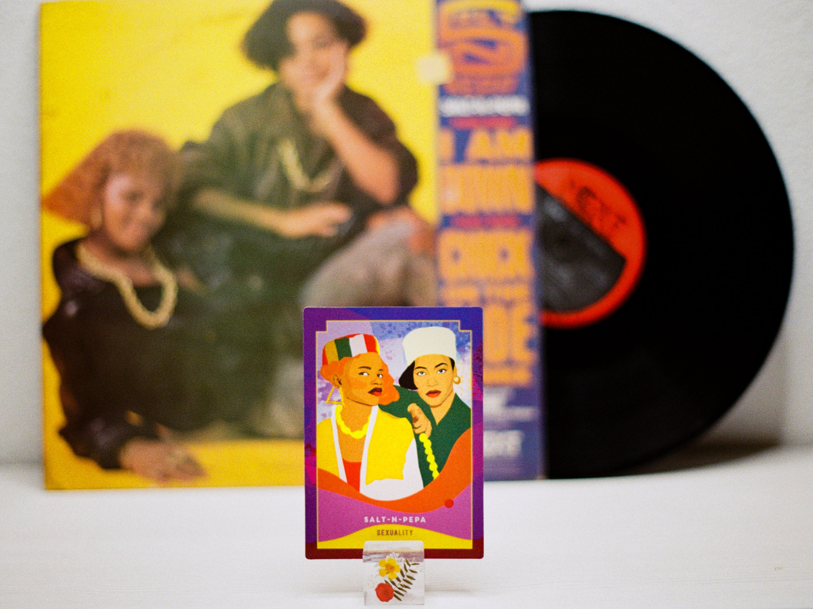 Vinyl record with a colorful cover featuring two people, a smaller card with a similar design, and a yellow album cover in the background.