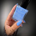 A hand with light blue manicured nails holds a bottle of Retrograde Rescue nail polish. The nail polish inside the clear glass bottle is a light sky blue, and the bottle has a black cap.