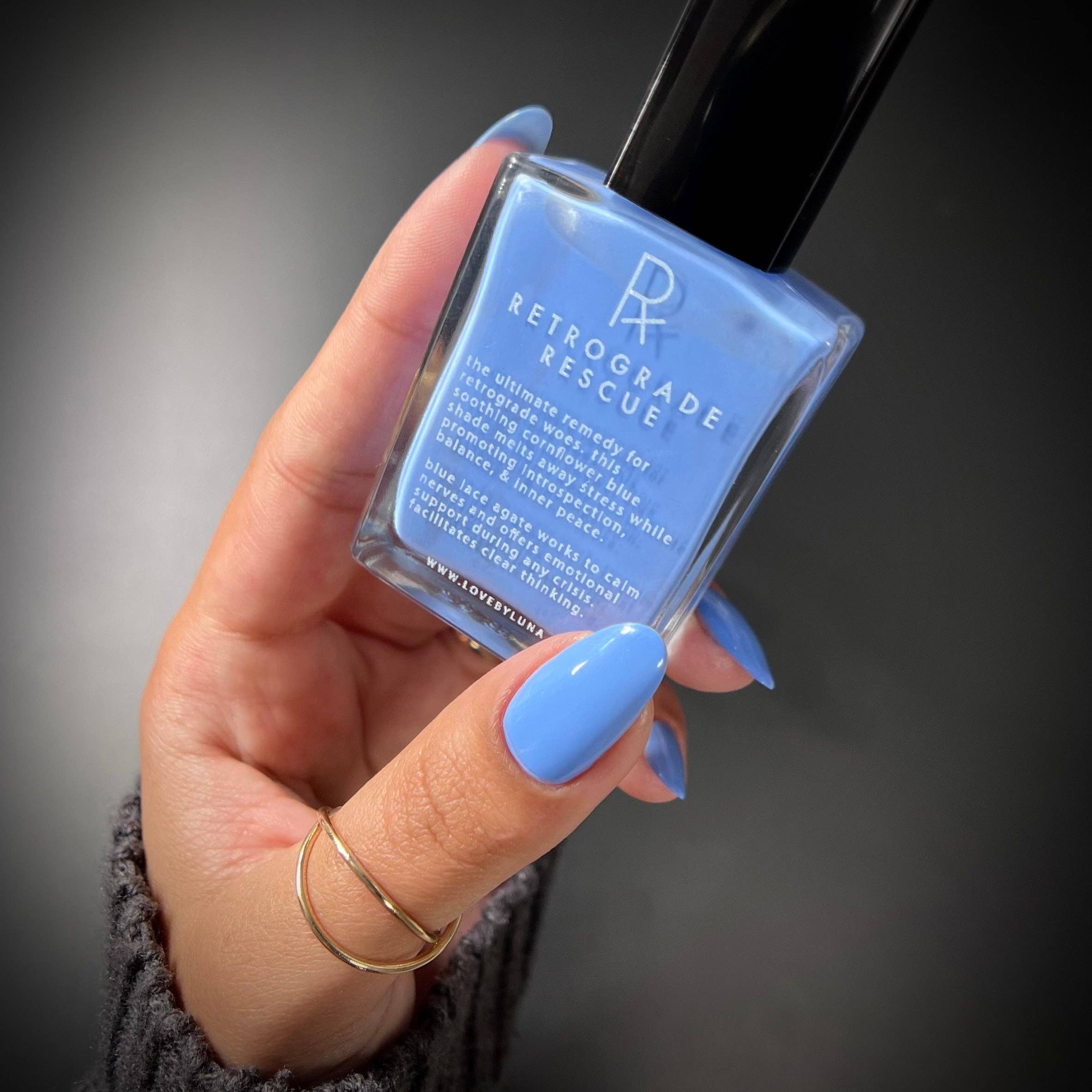 A hand with light blue manicured nails holds a bottle of Retrograde Rescue nail polish. The nail polish inside the clear glass bottle is a light sky blue, and the bottle has a black cap.